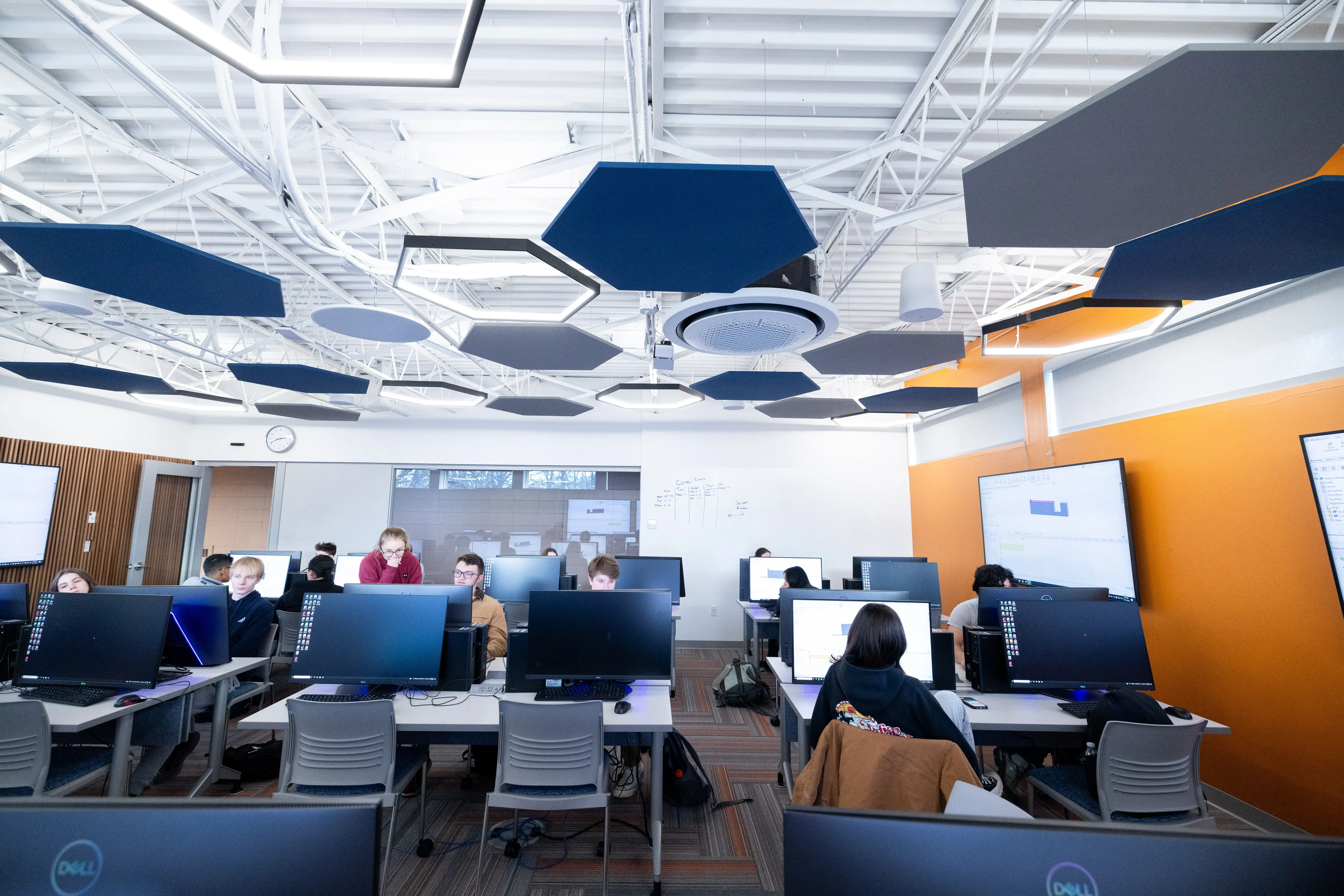 Students work at desktop monitors in a bright, modern computer lab with orange walls, whiteboards, and large display screens.