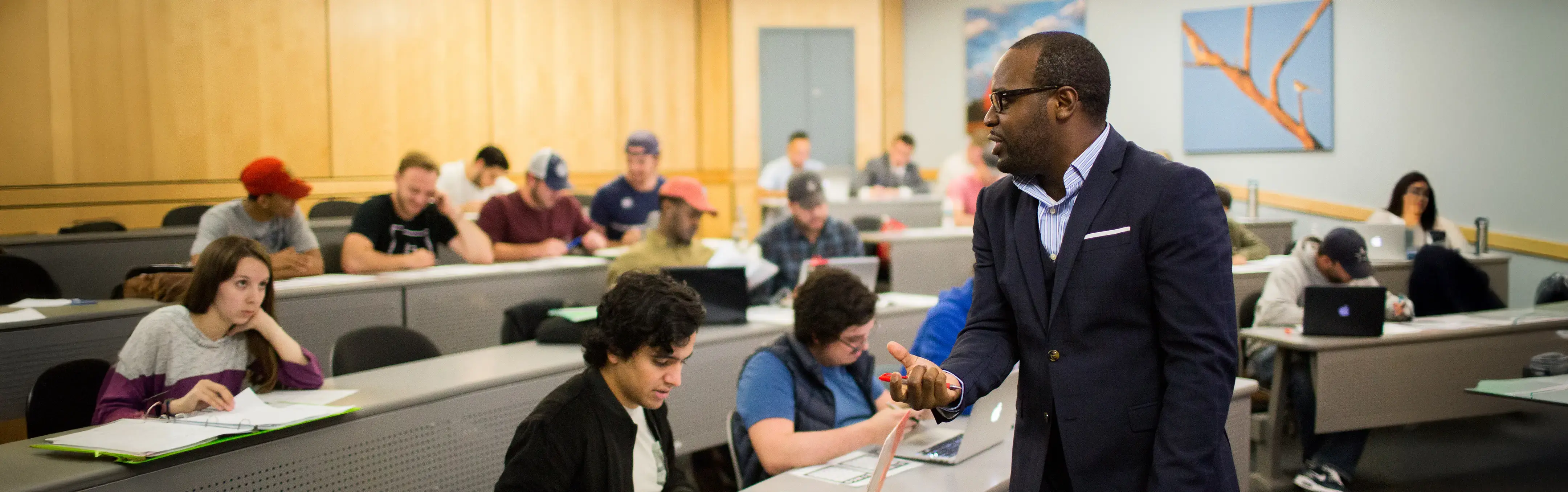 A professor in a suit assists students working on laptops and papers in an accounting classroom.